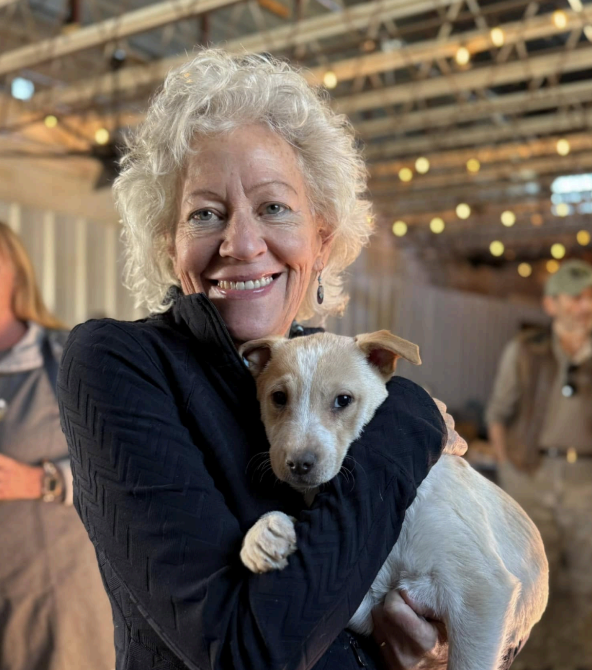 Woman with curly gray hair smiles, holding a puppy. Indoor setting with string lights and other people.