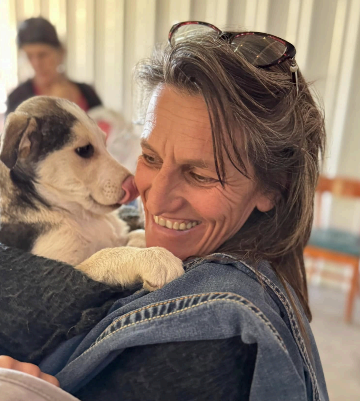 Woman smiles, holding a puppy, both looking at each other. Indoors, another person in the background.