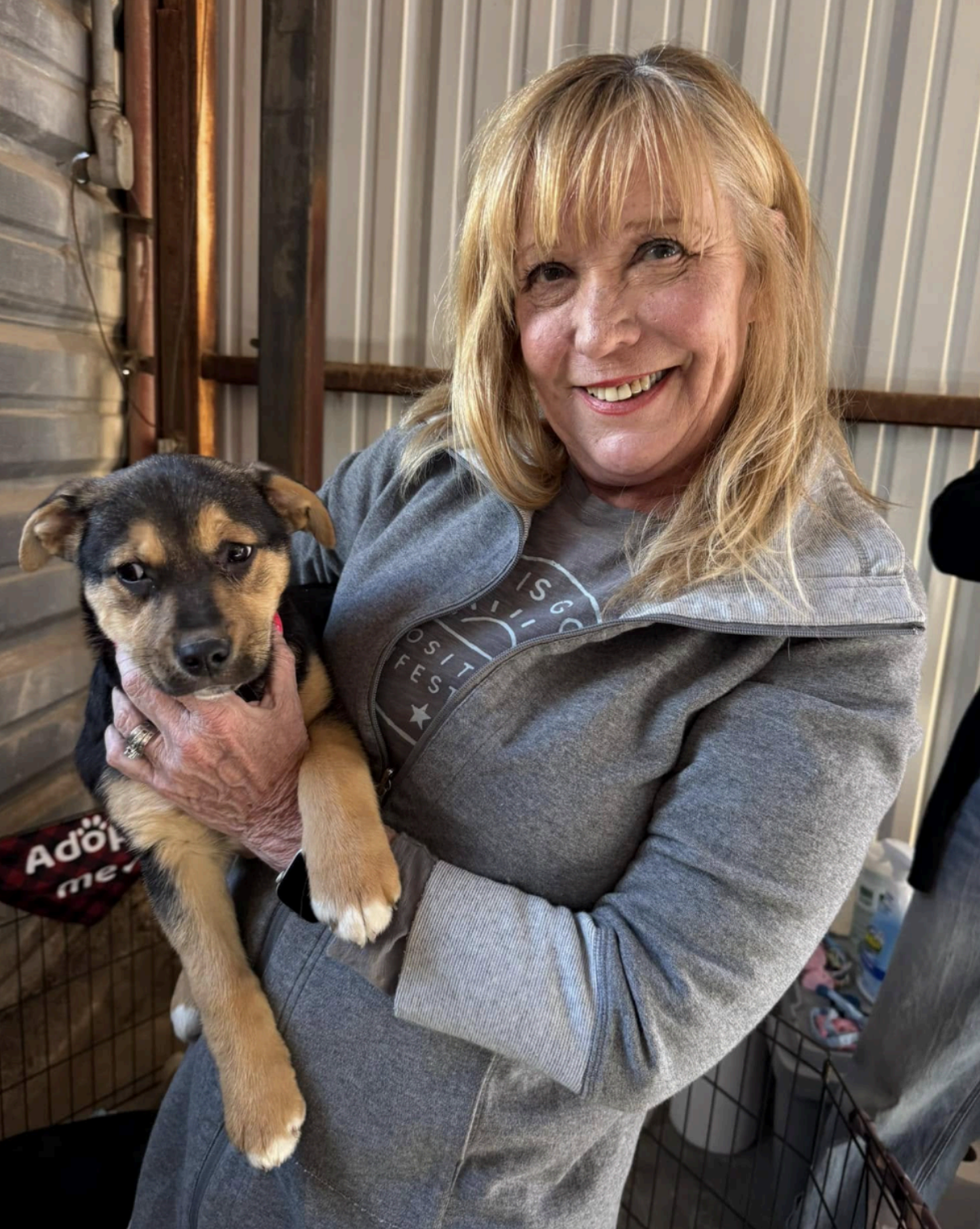 Woman smiles, holds a puppy. Puppy has black and brown fur. Shelter setting with corrugated metal walls.
