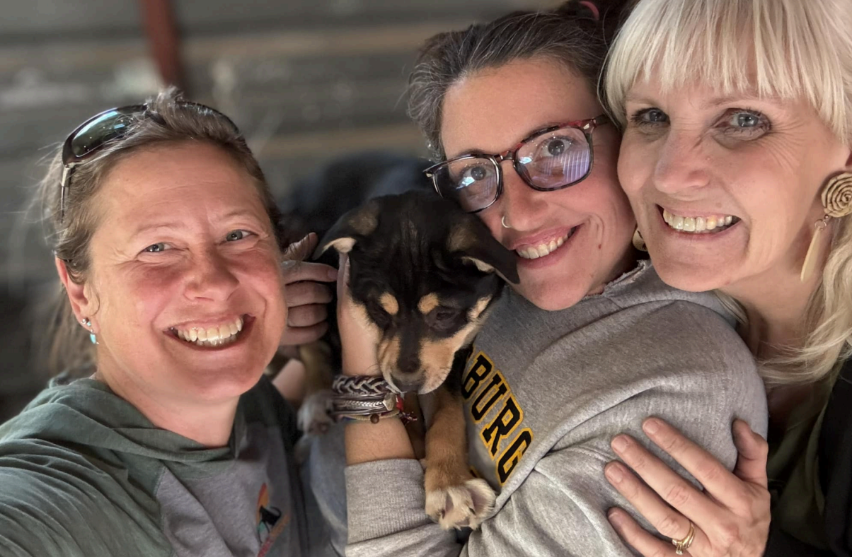 Three smiling people, holding a small dog. One woman in glasses hugs the dog, two others are on either side.