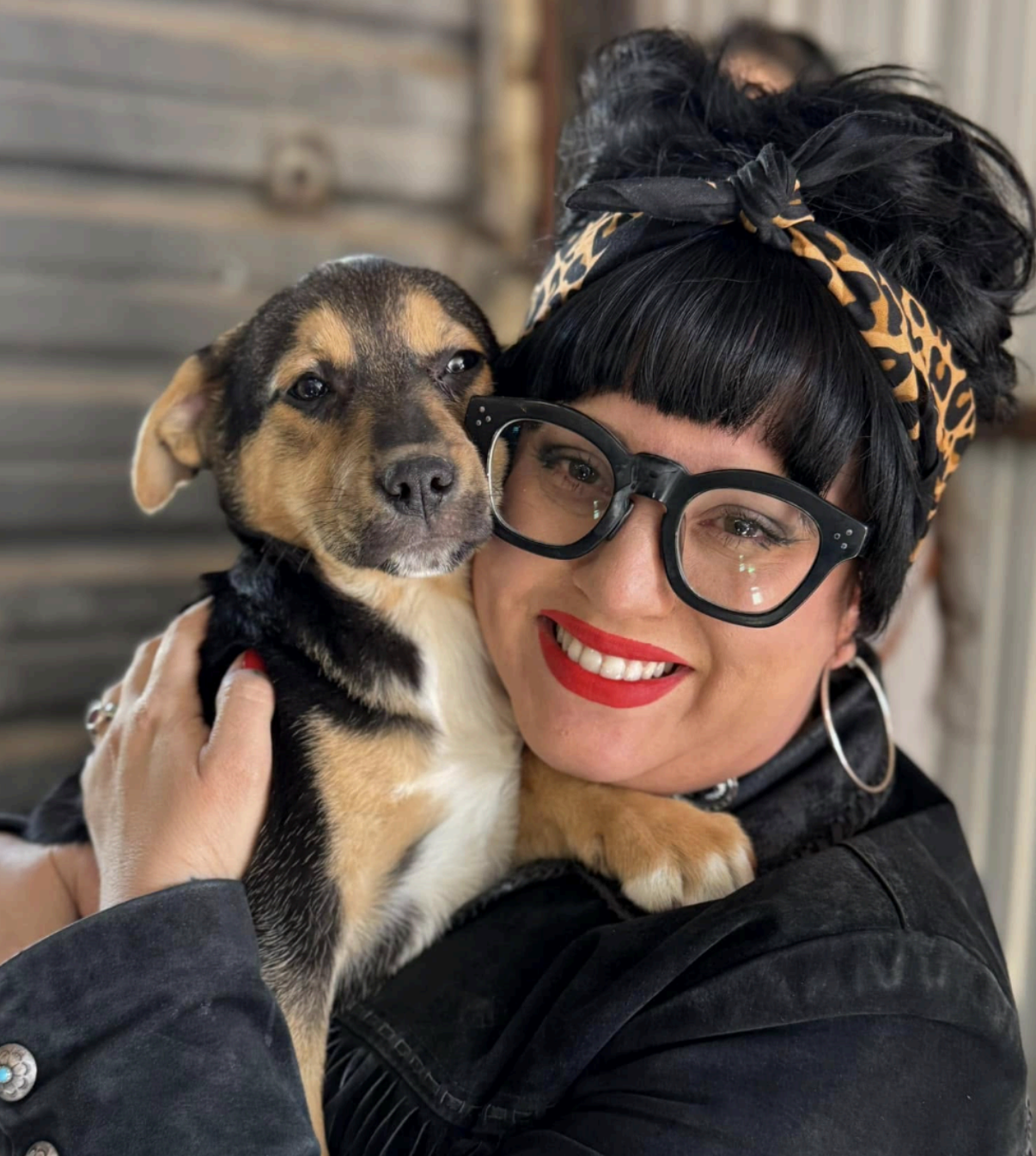 Woman in glasses hugs a puppy, smiles. They are outdoors near a rustic building.