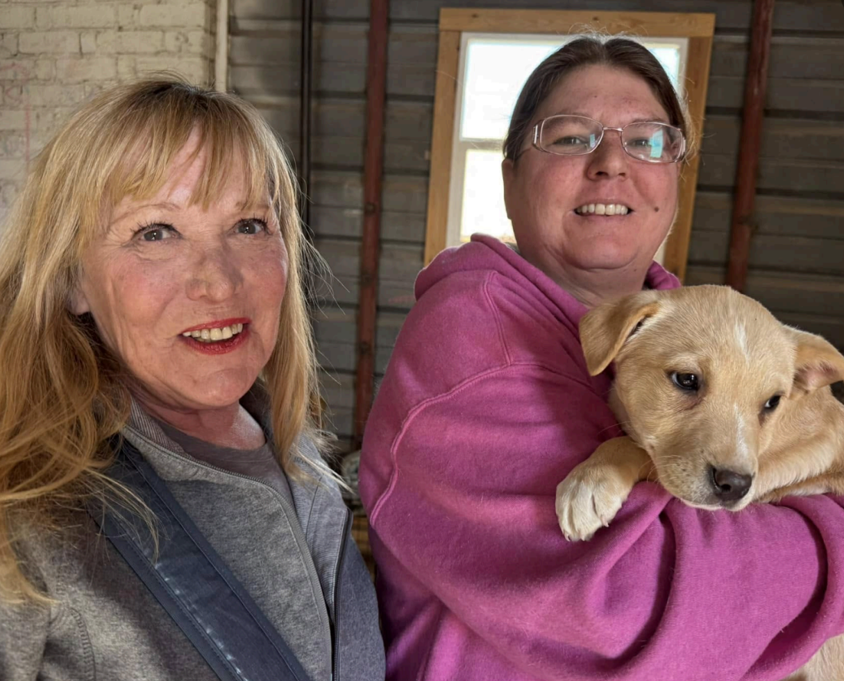 Two women with a puppy. One holds the tan pup in a pink hoodie. Blonde woman smiles. Building interior.