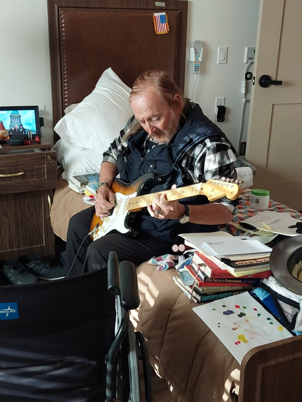 Man playing a guitar while seated on a bed in a room, stacks of paper beside him.