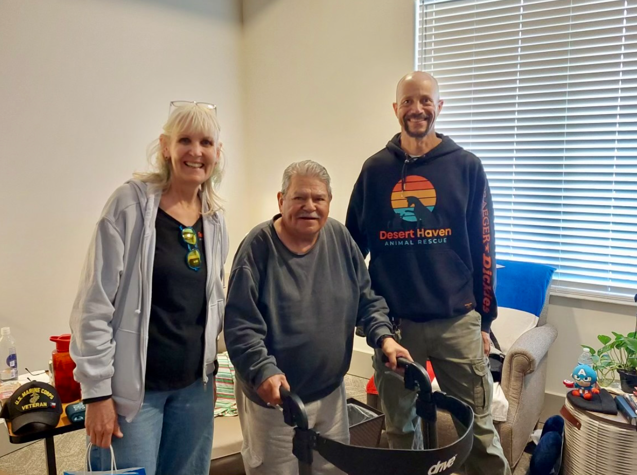 Three people pose indoors; one uses a walker. Two on either side smile.