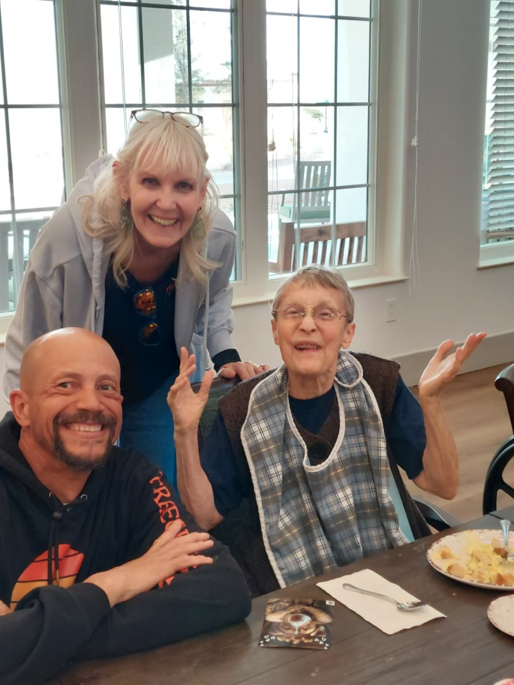 Three people smiling at a table. One with a bib and arms raised. One is smiling behind them.