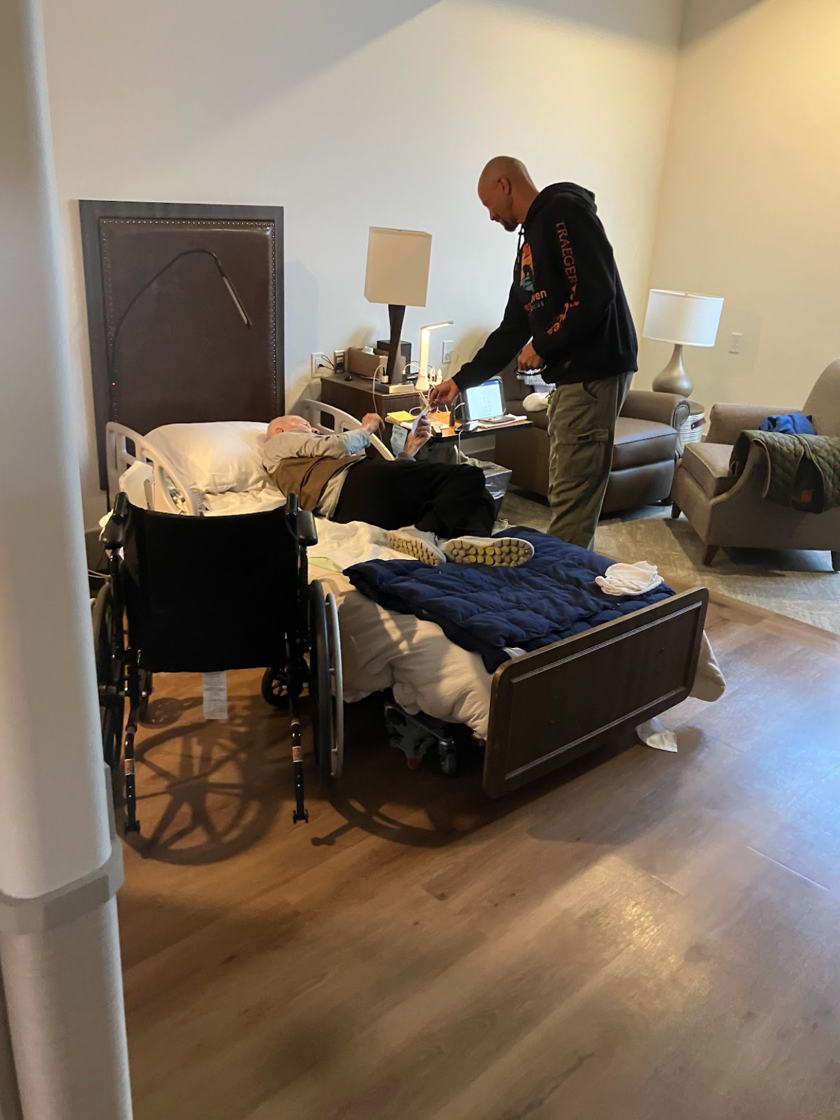 A person tends to a hospital bed next to a wheelchair. Interior room with lamps and furniture.