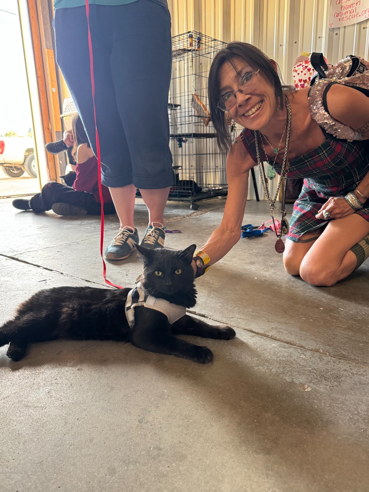 Woman kneels to pet a black cat on a leash inside a building. Another person stands nearby.
