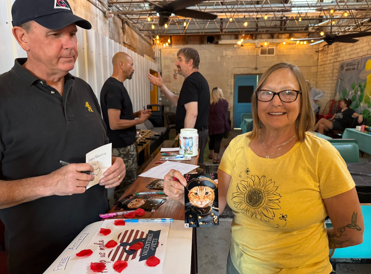 People at an event. A woman smiles next to a table with signed artwork. A man in a cap holds a pen.