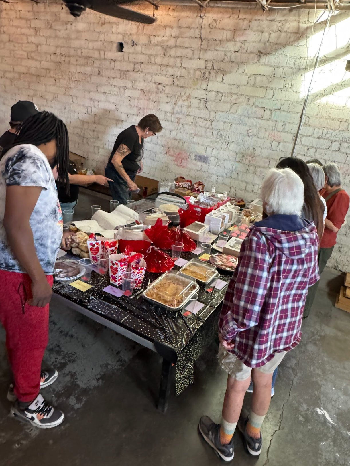 People browsing a table of food. A brick-walled room. Various desserts in containers.