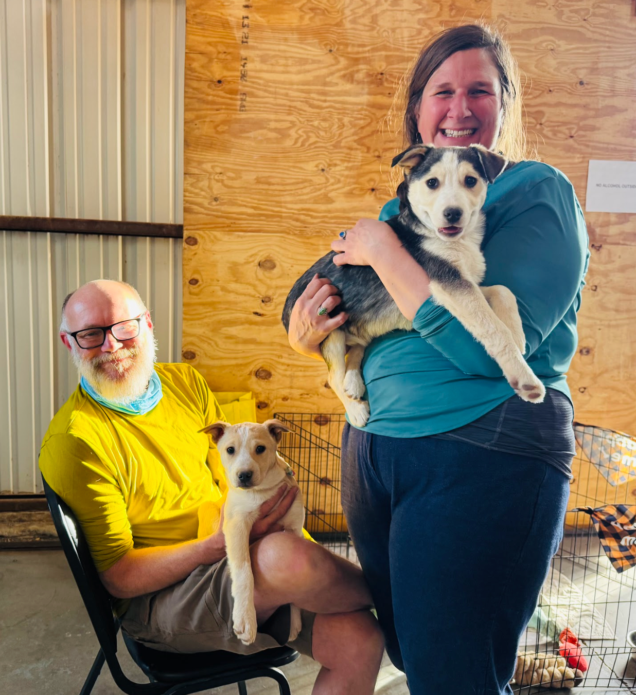 Two people with two dogs in an indoor setting. One woman holds a puppy, and a man sits holding another.