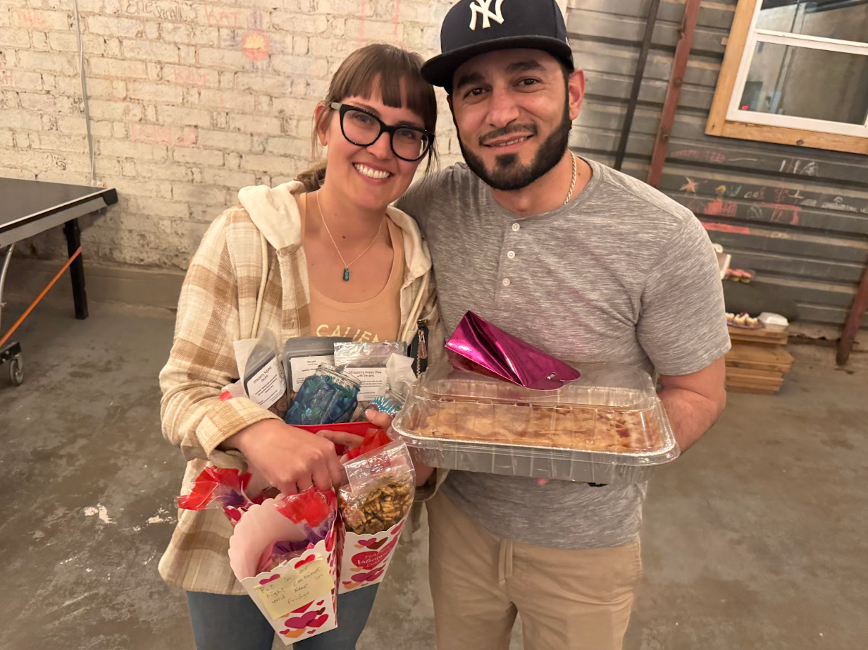 Smiling couple holding gifts and a cake in a brick-walled room.