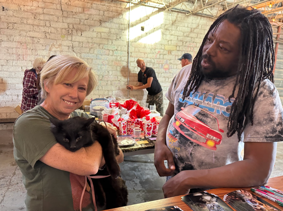 Woman holding black cat, smiling at man with dreadlocks. Others work on wall in a brick-walled space.