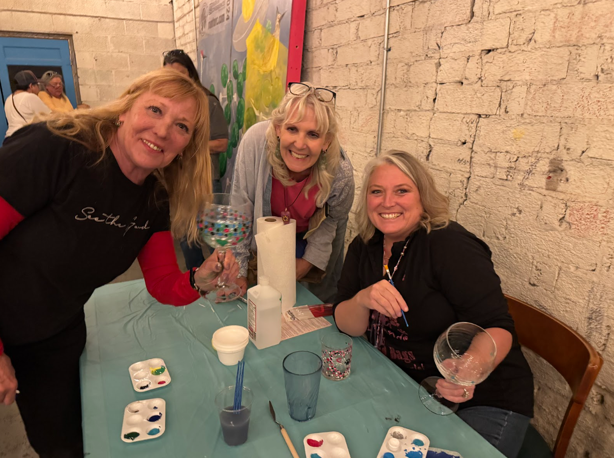 Three women smile at a table with painting supplies, holding wine glasses, in a studio.