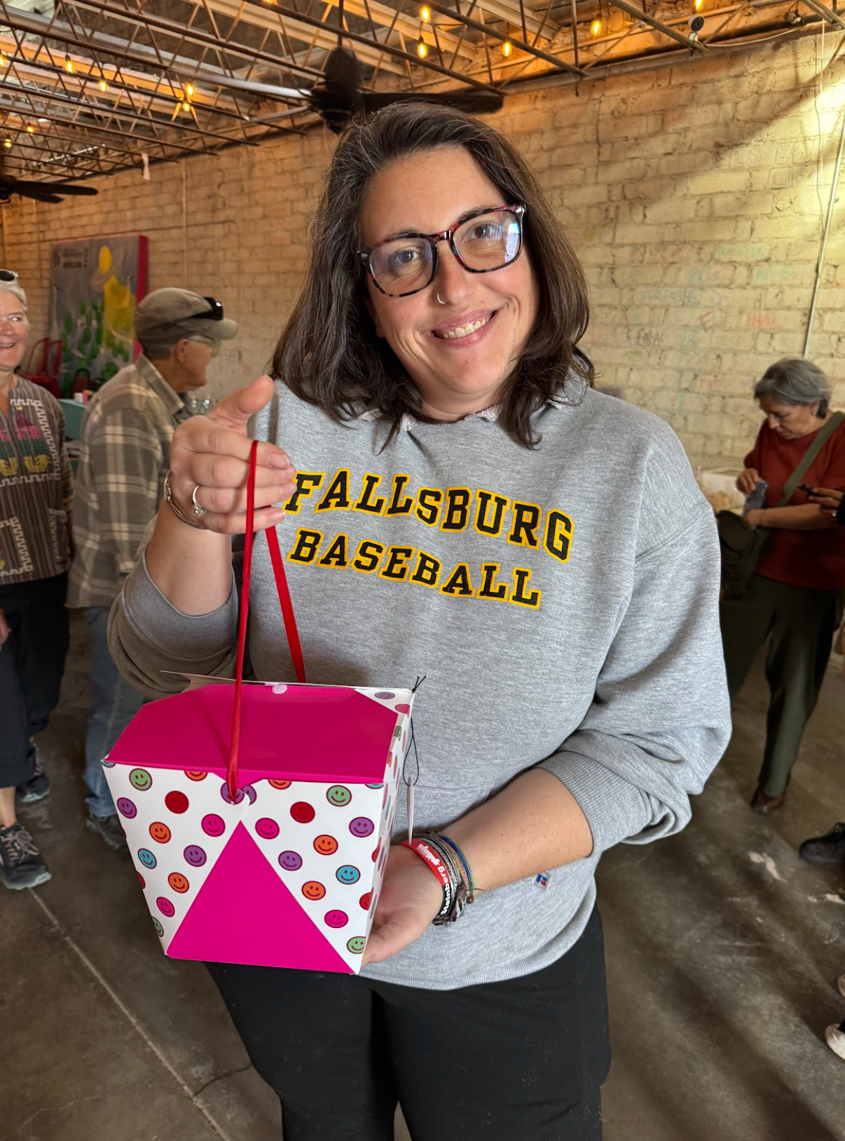 Woman holding a handmade pink and polka-dot purse, smiling. Wearing glasses and sweatshirt. Inside a room with people.