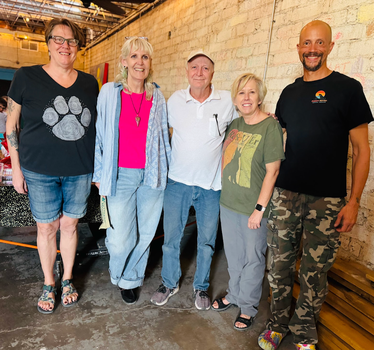 Five people standing together, smiling, against a brick wall. People are wearing casual clothing.