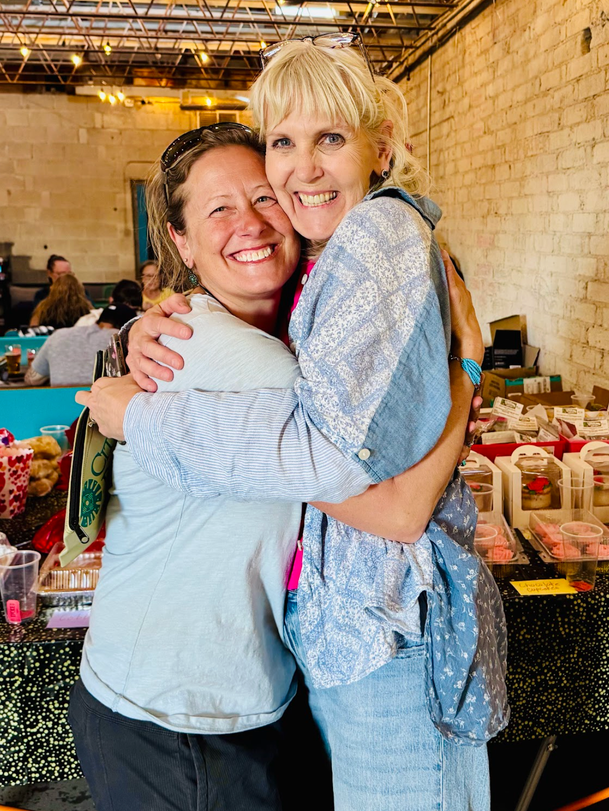 Two women embracing, smiling; outdoors market setting with baked goods, light blue clothing.