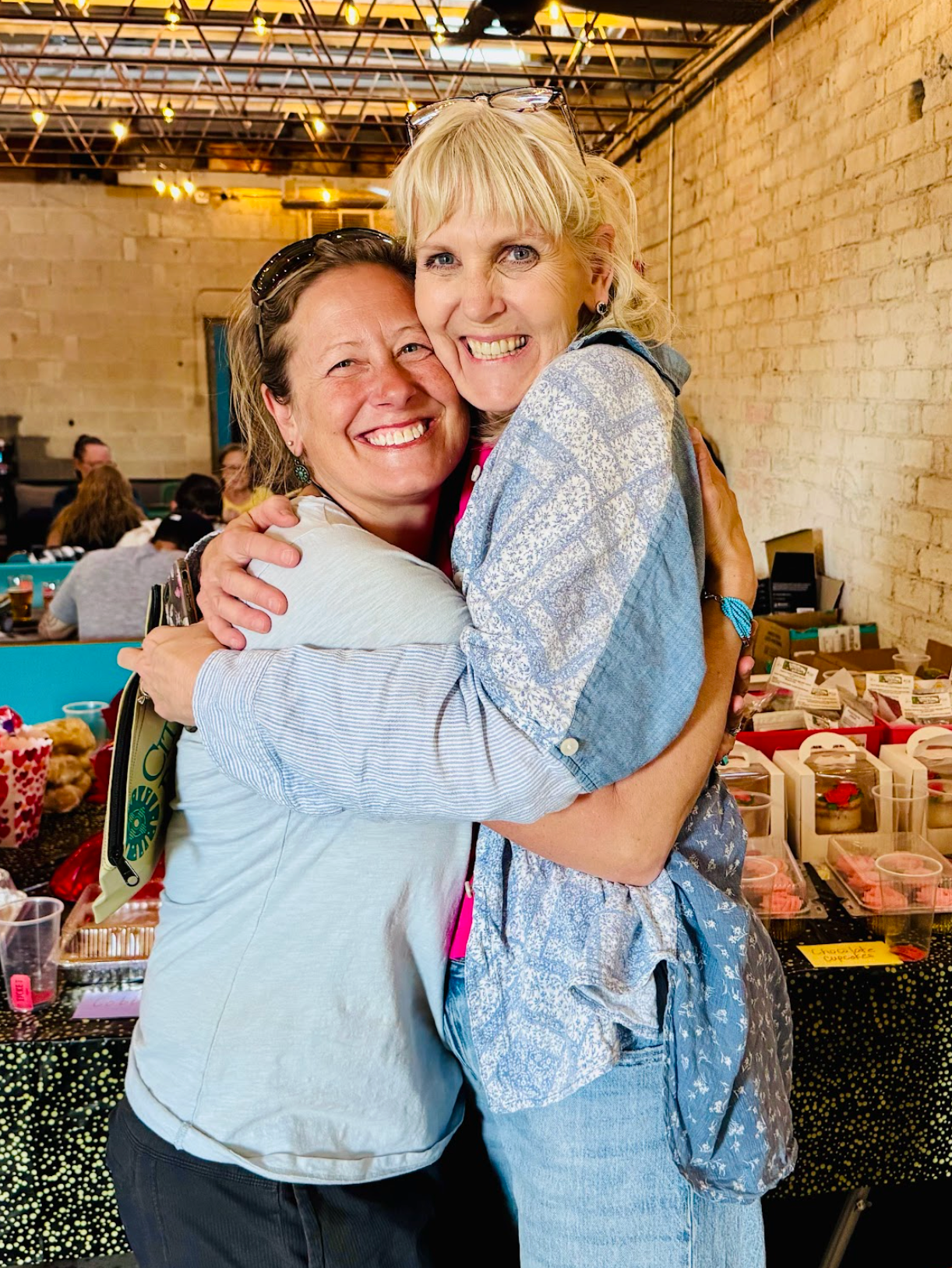 Two women embrace, smiling. Bright indoor market setting, tables in the background.