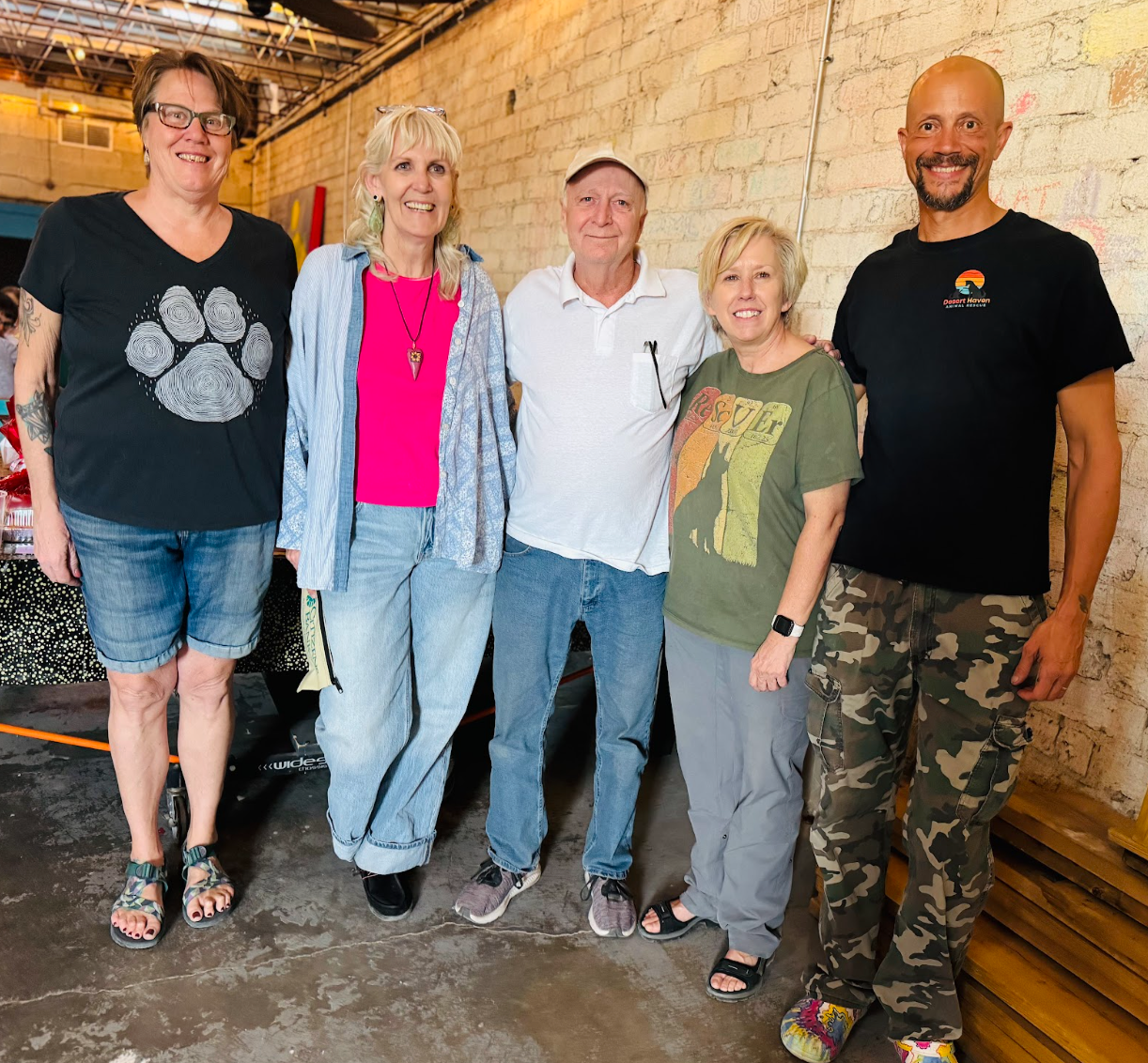 Five people standing, smiling, next to a brick wall. One wears a paw-print shirt, another pink.