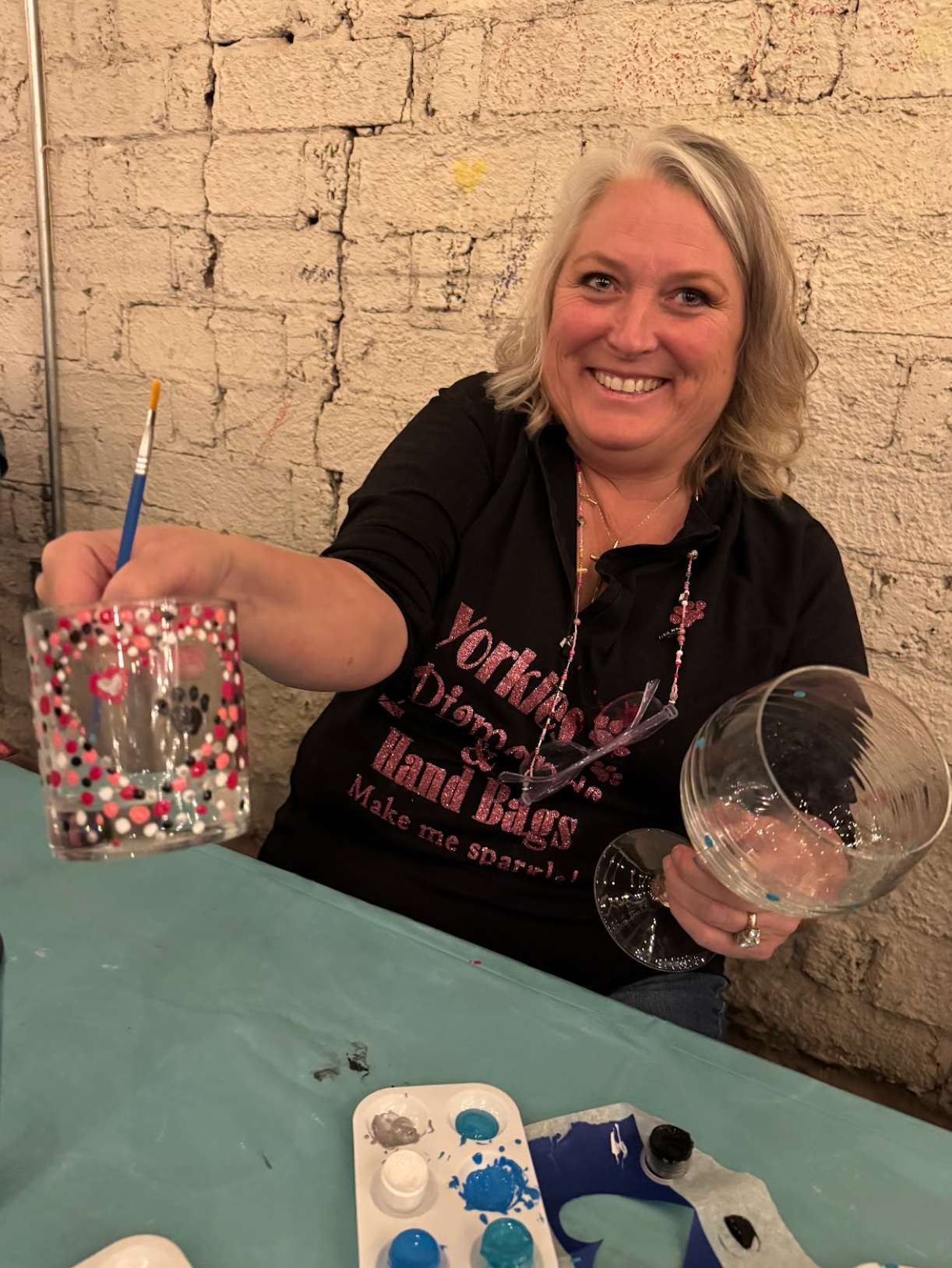Woman holding painted glass and paintbrush, smiling at painting class. White and pink heart design on glass.