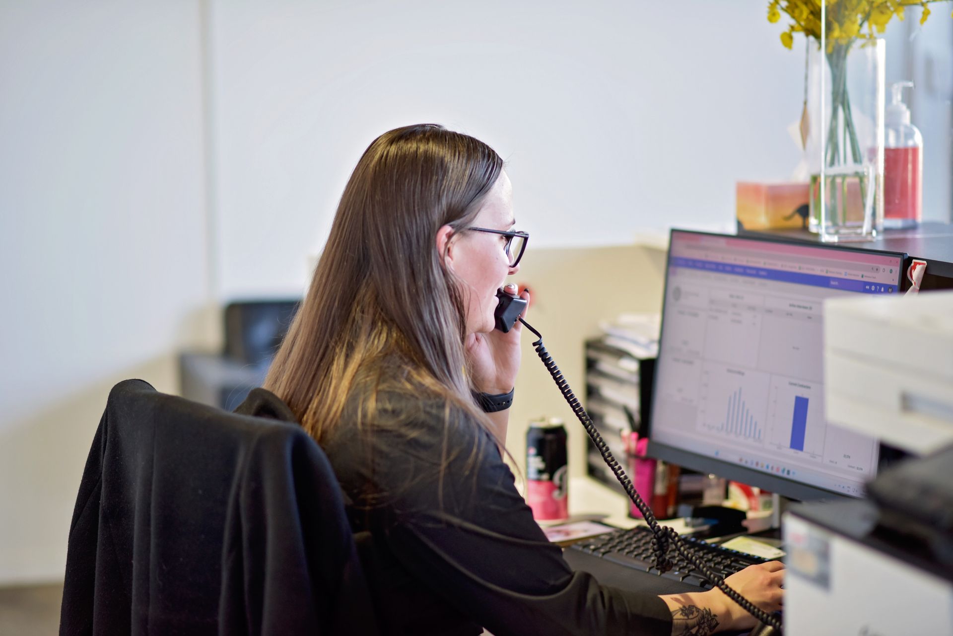 A woman is sitting at a desk talking on a phone in front of a computer.