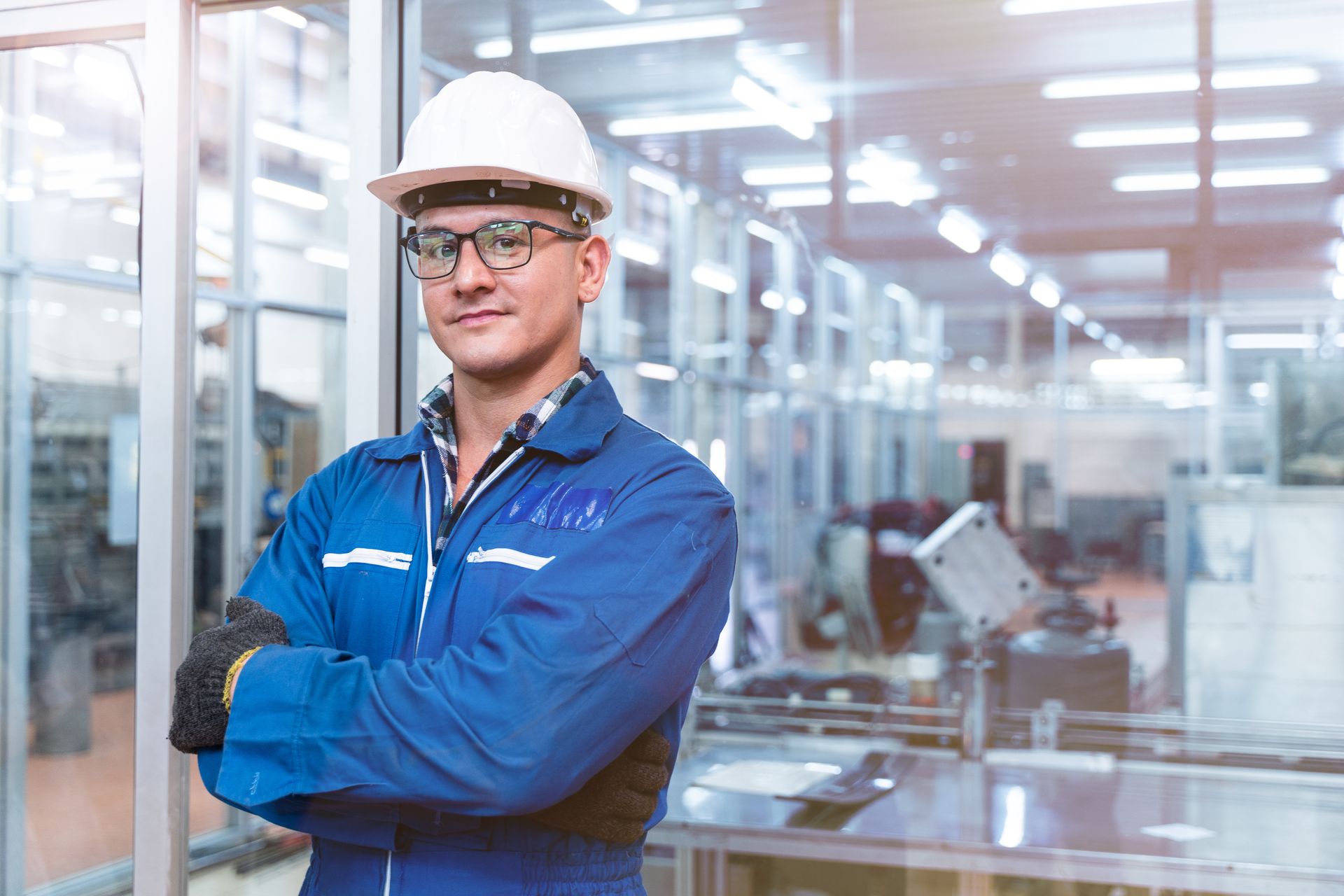 A man wearing a hard hat and safety glasses is standing in a factory with his arms crossed.