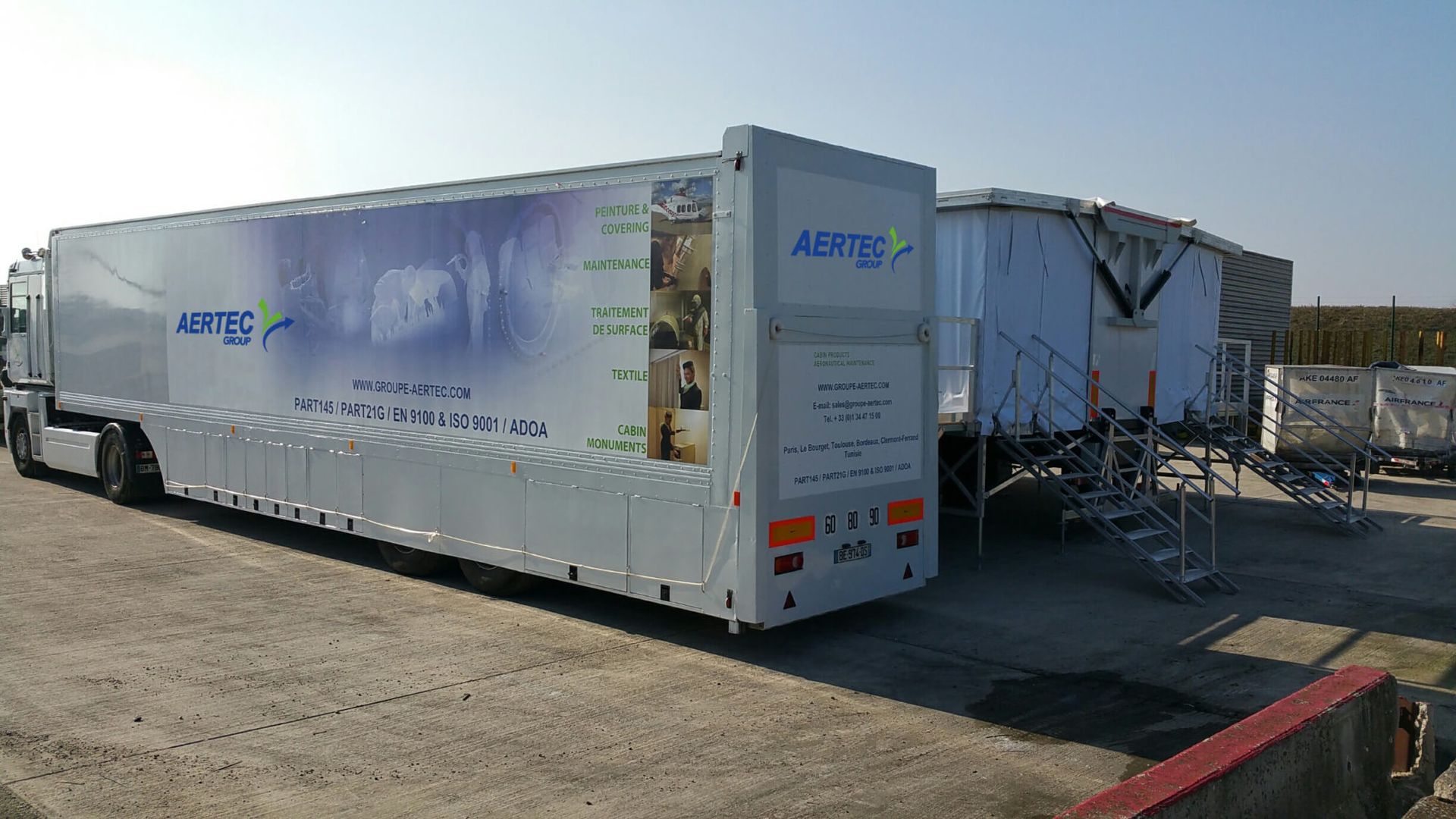 Semi-remorque blanche portant le logo « AERTEC ». Une autre structure se trouve à droite, sur une surface en béton.
