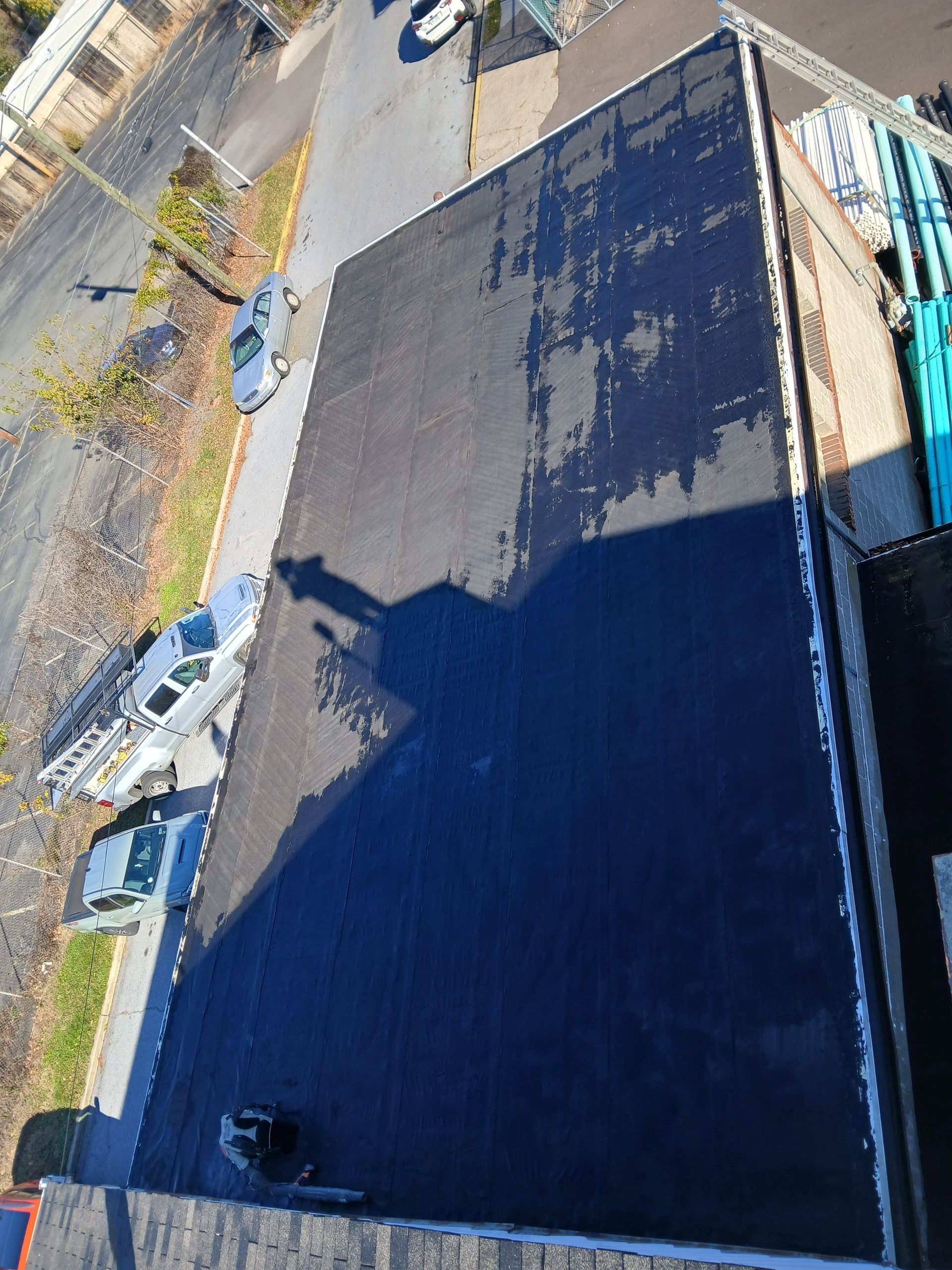 Aerial view of a large dark rooftop beside parked cars and a road lined with trees.