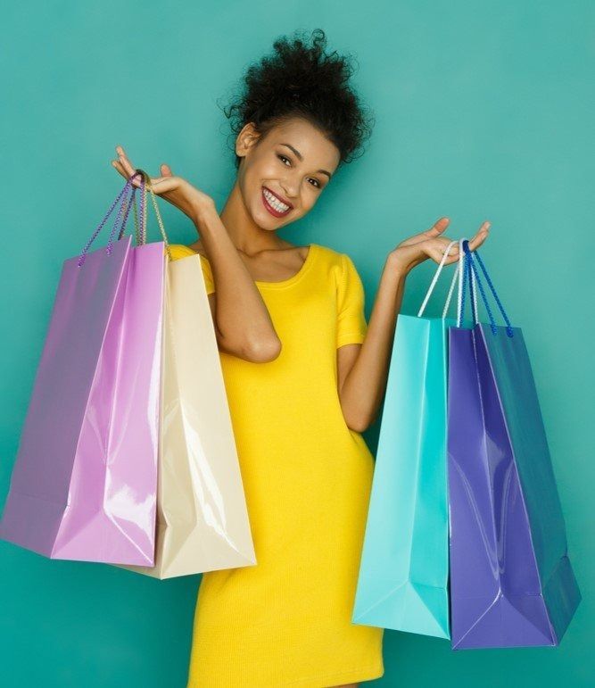 Smiling person in a yellow dress holding colorful shopping bags against a teal background.