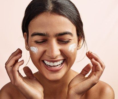 Smiling person applying white cream to both cheeks against a soft pink background