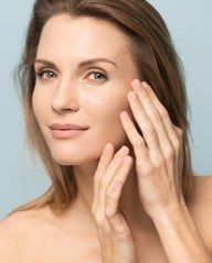 Woman gently applying skincare cream to her cheek against a light background