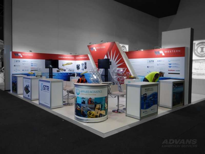 Trade show booth with white, red, and blue branding. Displays, tables, and a person working are visible.
