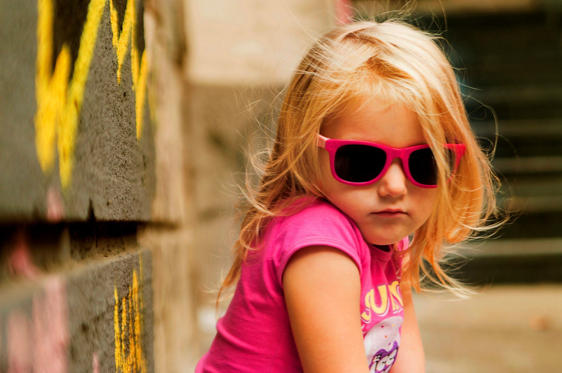 Bambino con occhiali da sole e maglietta rosa appoggiato a un muro all'aperto.