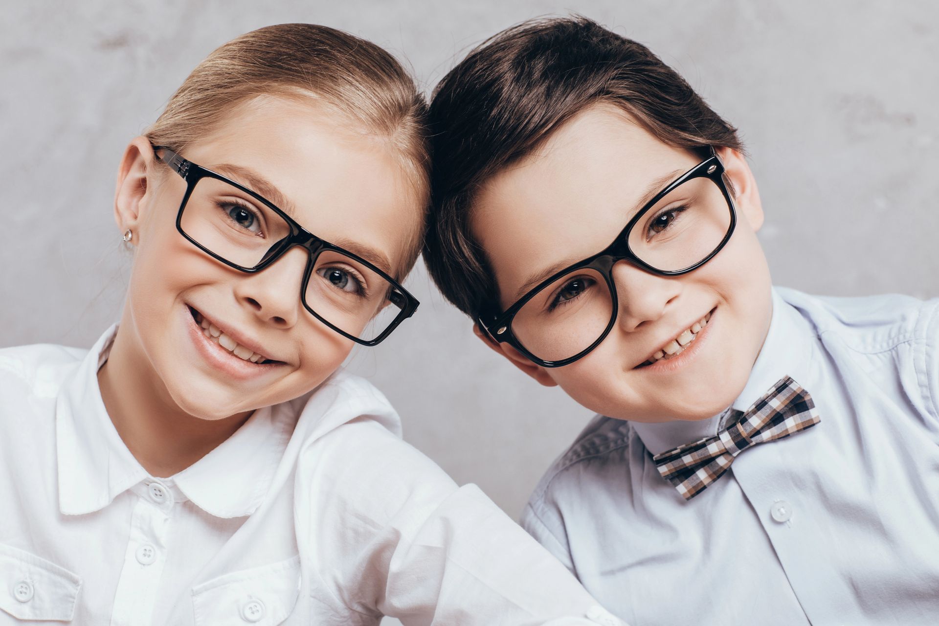Due bambini con camicie bianche e occhiali sorridono su uno sfondo grigio chiaro.