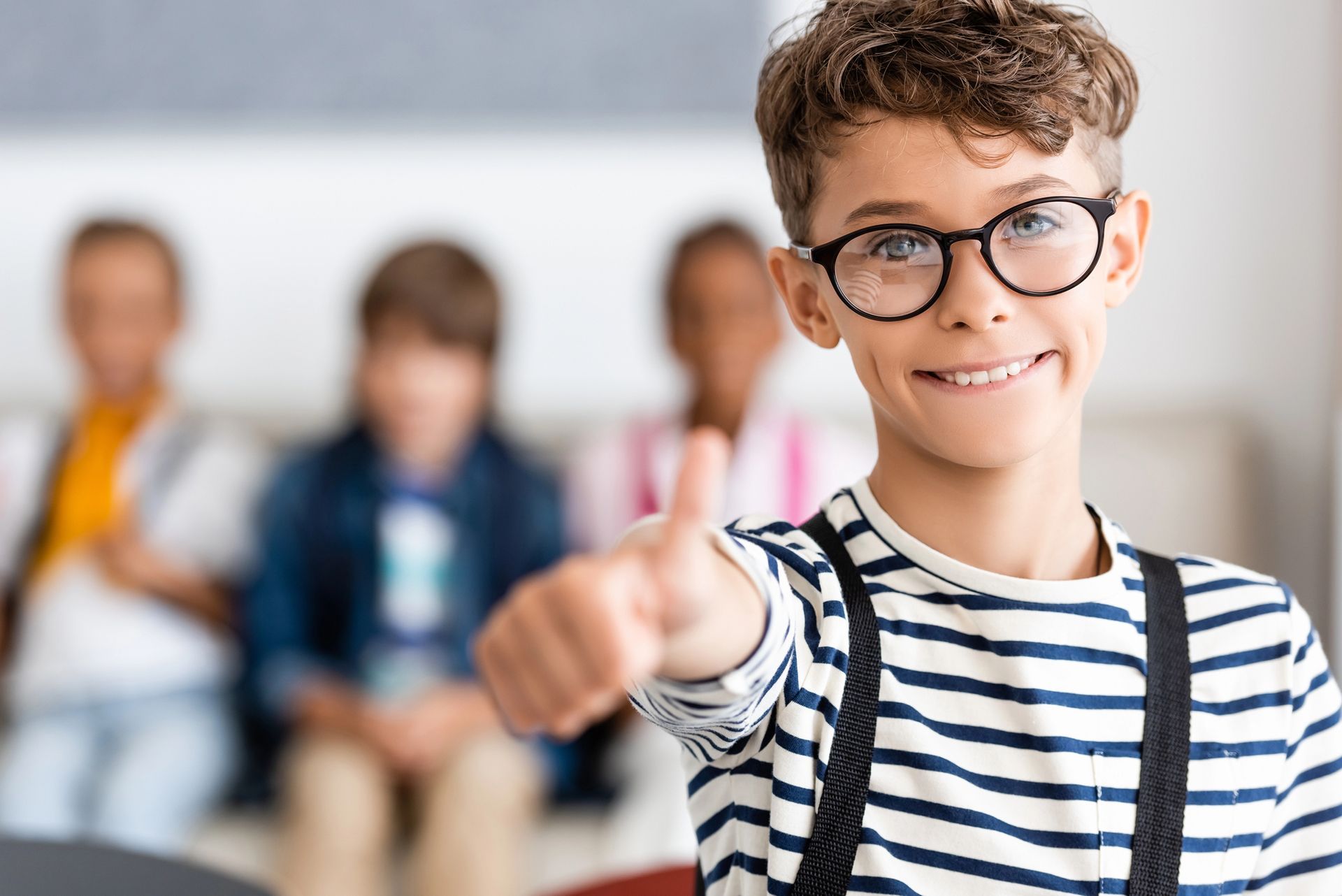 Bambino sorridente con occhiali e maglietta a righe che fa il segno del pollice in su in un'aula scolastica.