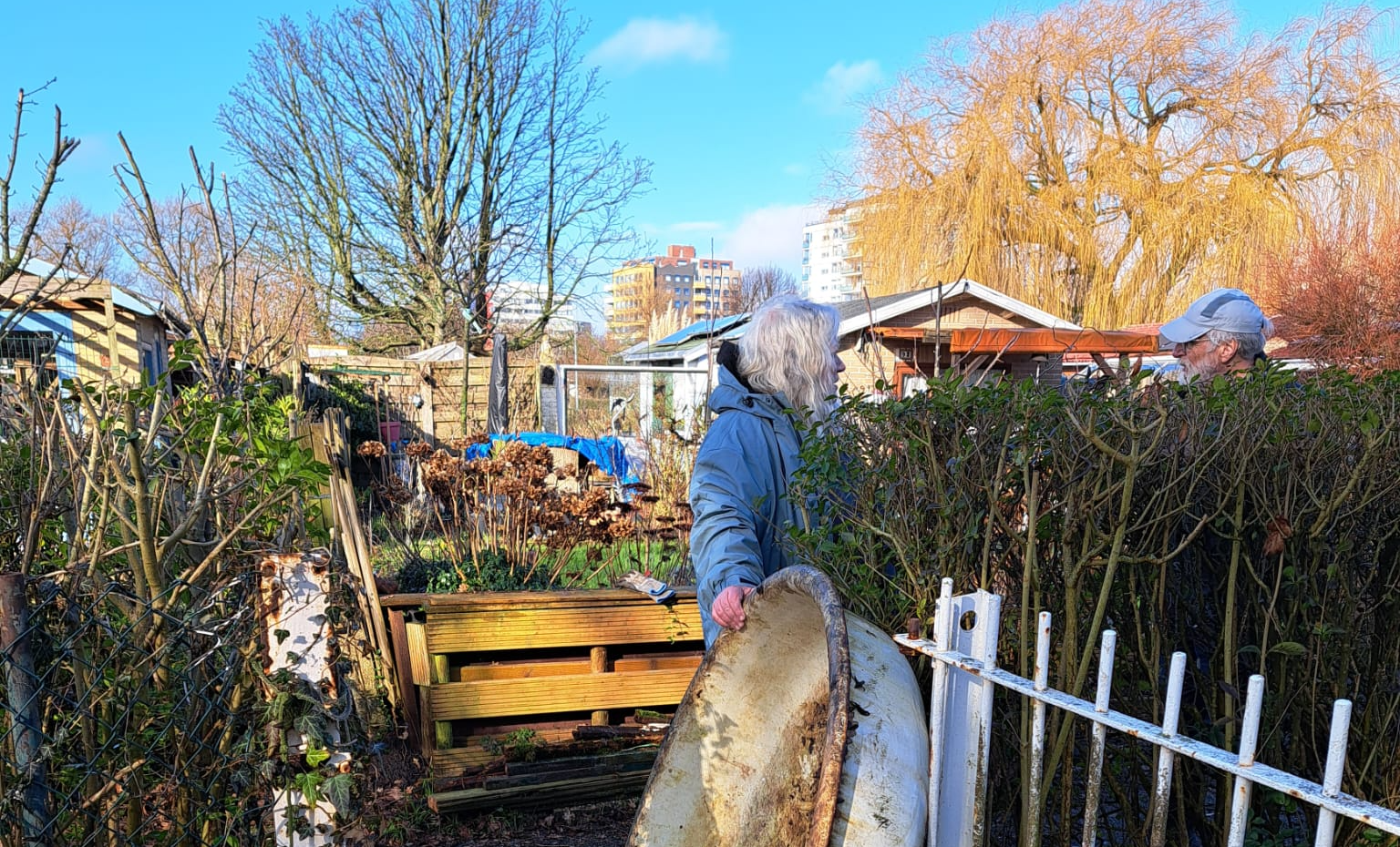 Een man draagt ​​een metalen voorwerp door een poort in een tuin; een andere persoon is zichtbaar bij een heg op een zonnige dag.