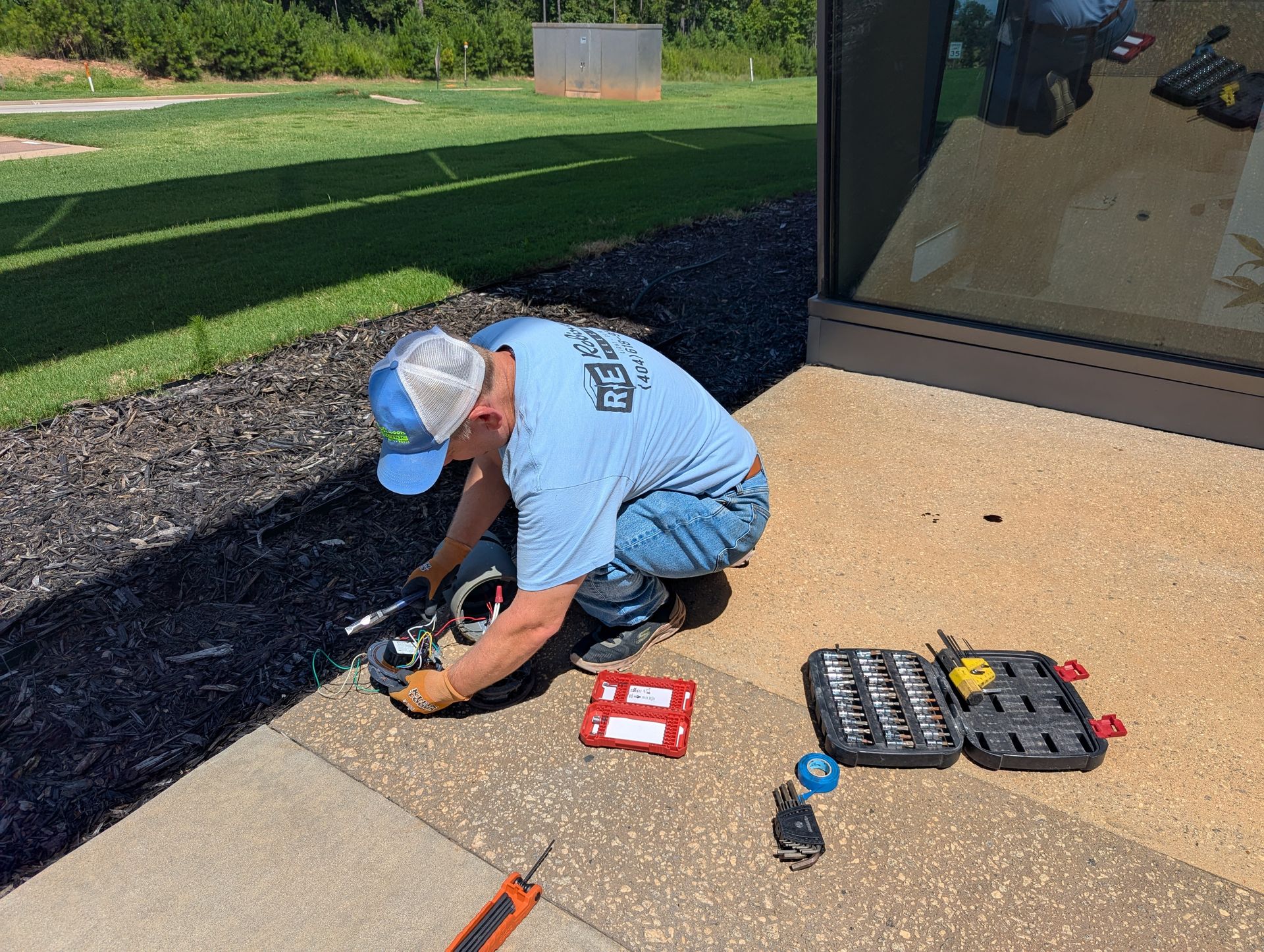 Man crouches, working on something near a building. Tools and a toolbox sit nearby on the ground.