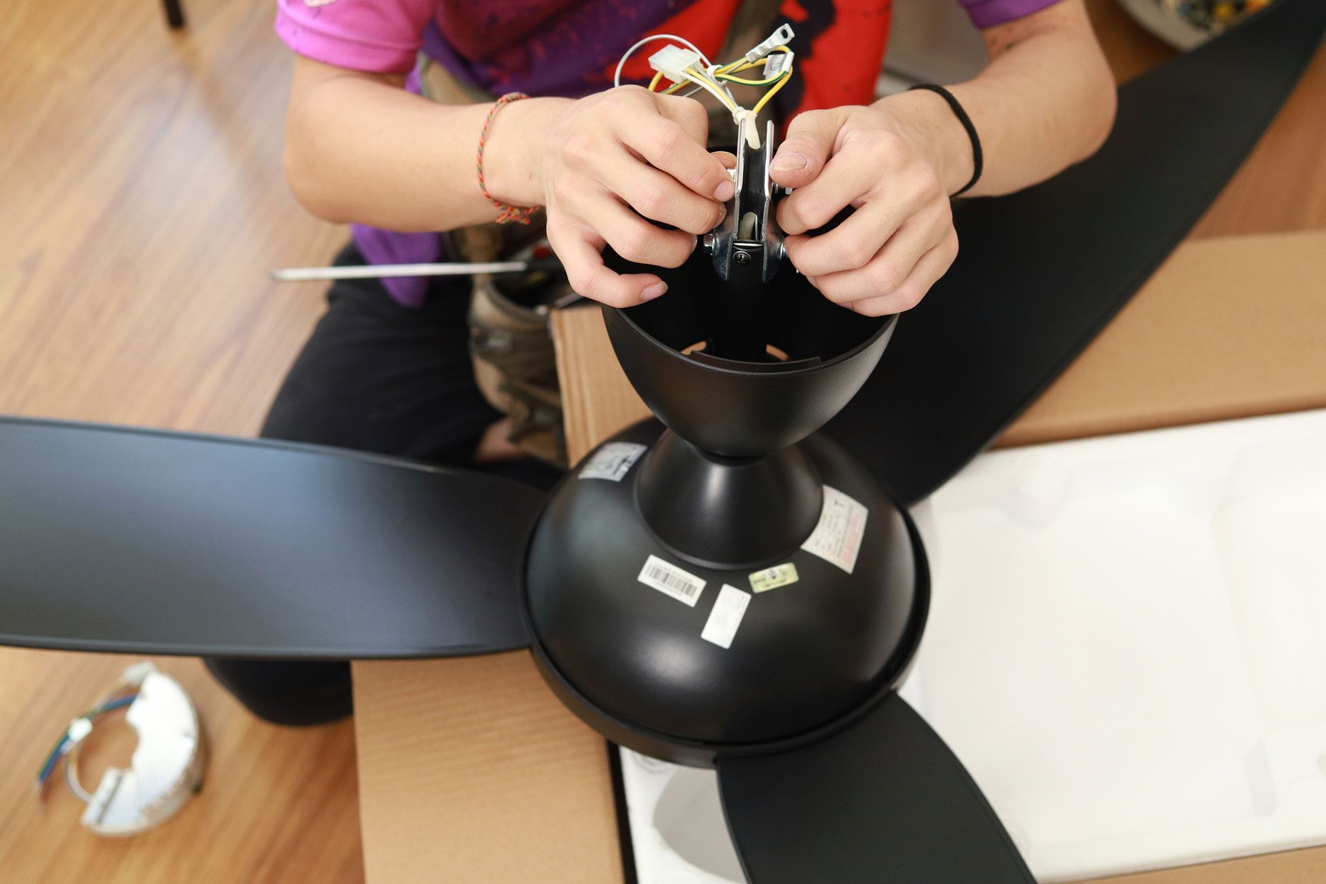 A person in a purple shirt assembles a matte black ceiling fan, connecting wiring to the motor housing above the blades.