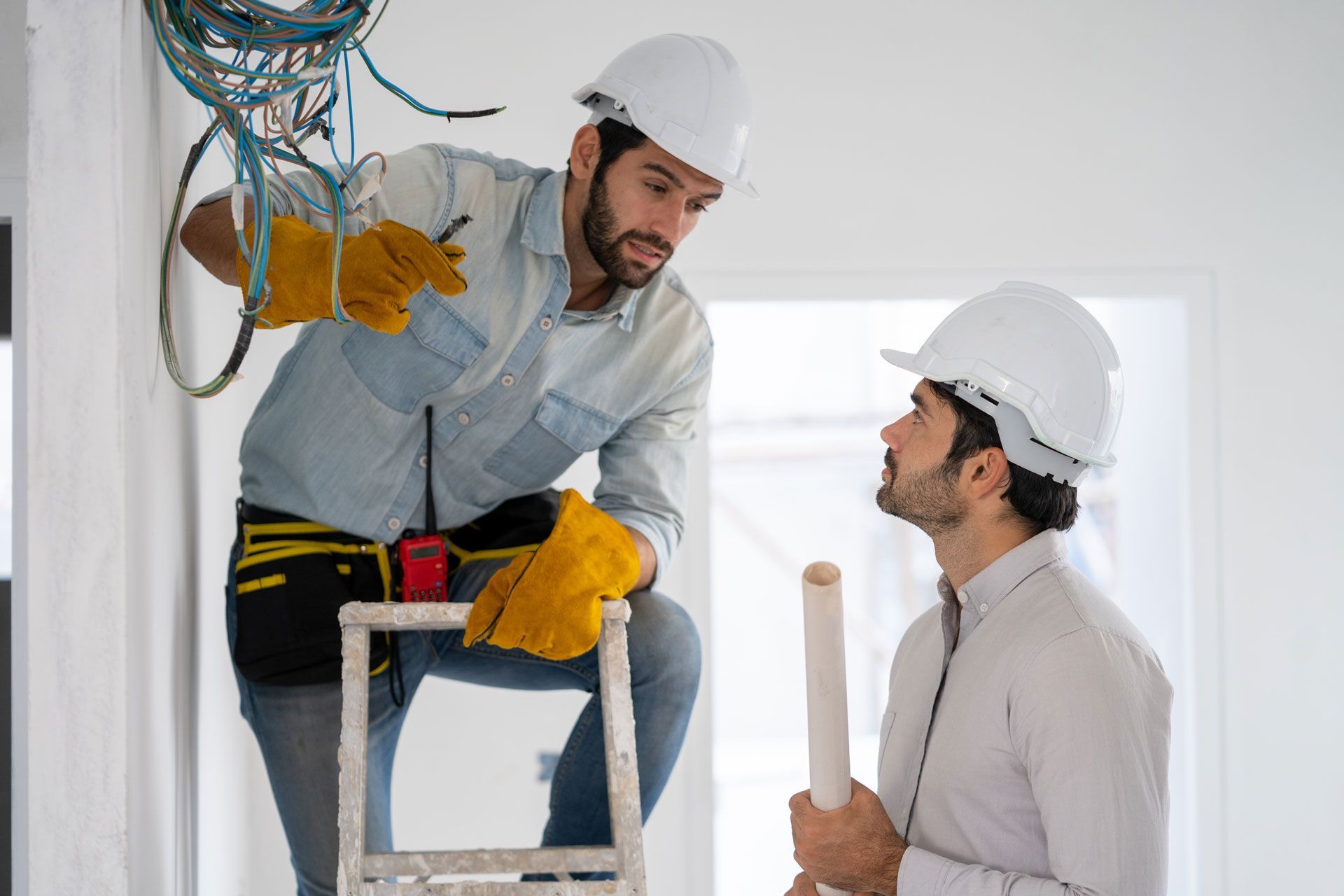 Two construction workers in hard hats discuss electrical wiring hanging from a ceiling inside a building.