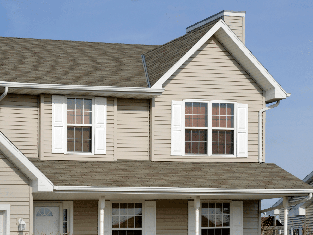Two-story beige house with white trim and shuttered windows under a blue sky