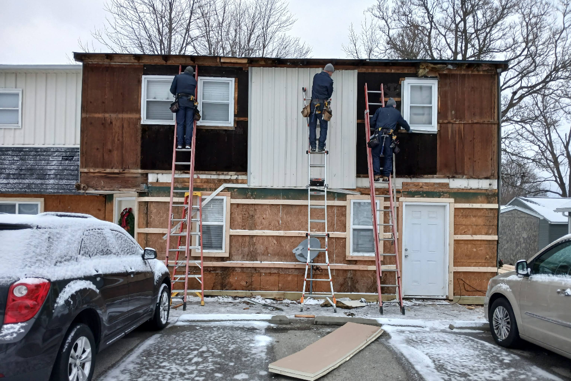 Workers on ladders repainting a two-story house with snow on the ground.