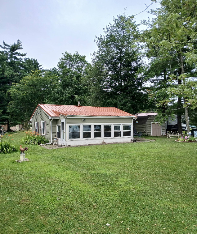 Small stone cottage with a red roof and front windows, set in a grassy yard with tall trees.