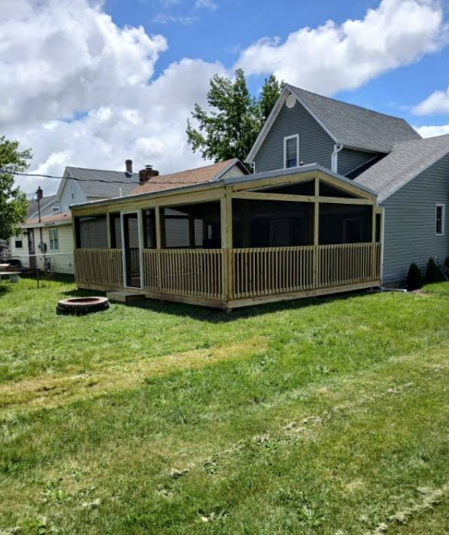 Gray house with a covered wooden porch and deck in a grassy yard under a partly cloudy sky