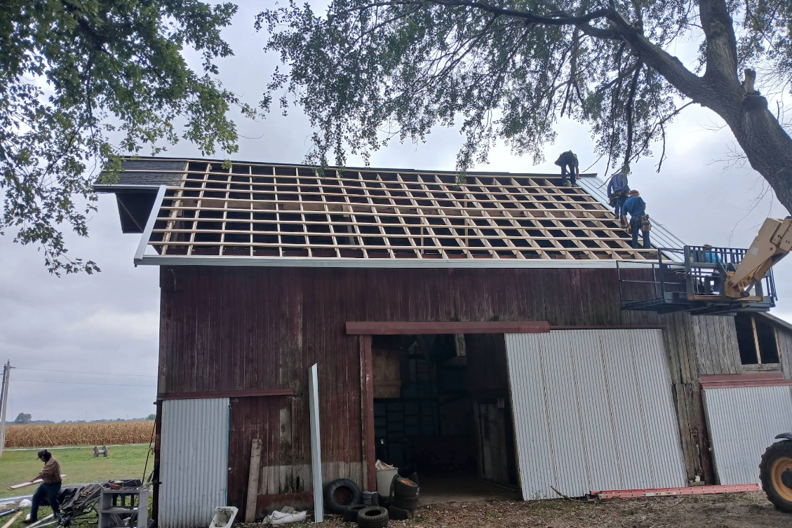 Workers reroofing a small barn with exposed rafters and metal siding in a rural field