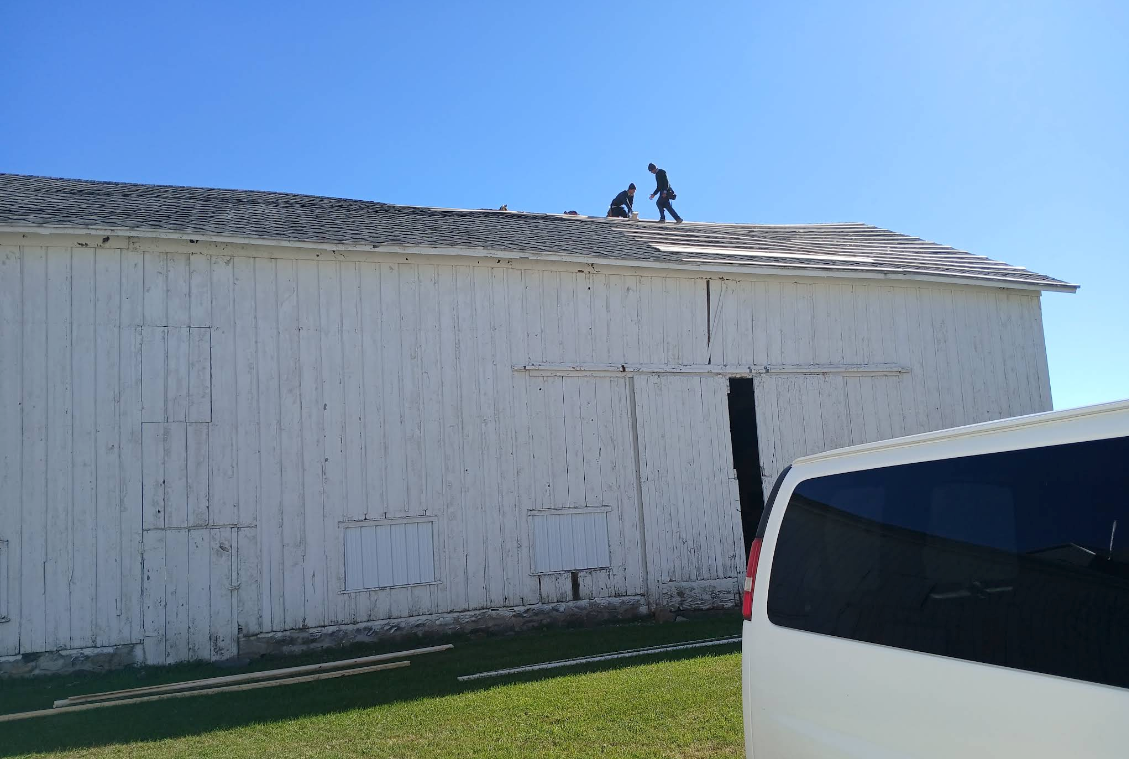 White building with damaged roof and two people standing on top, seen beside a white van on grass.