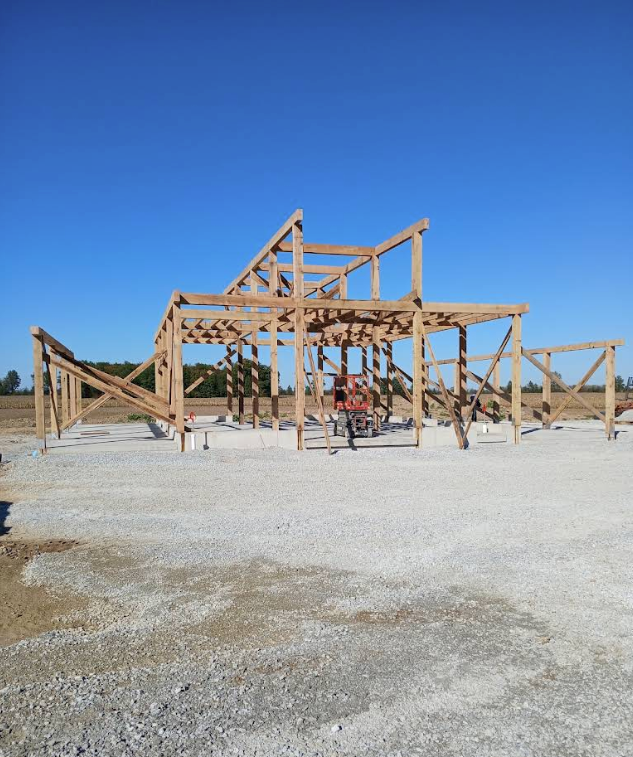 Wooden pavilion frame under construction on a gravel lot beneath a clear blue sky
