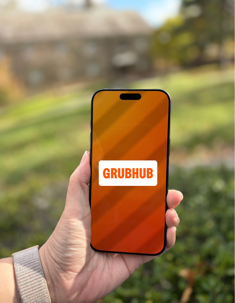 Hand holding phone displaying Grubhub logo. Outdoor setting with green grass and a building.