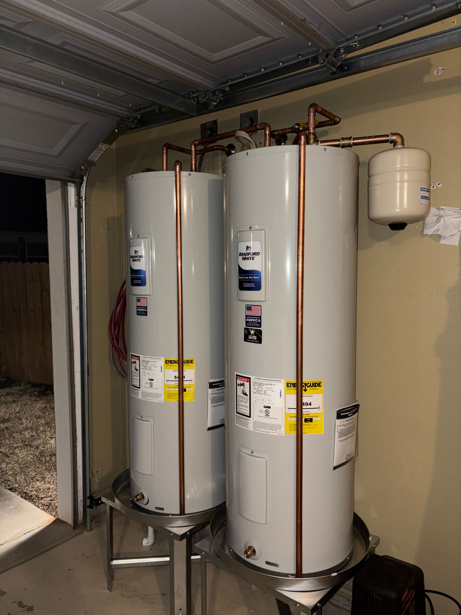Two tall white water heaters in a garage, with pipes and an expansion tank on the wall.