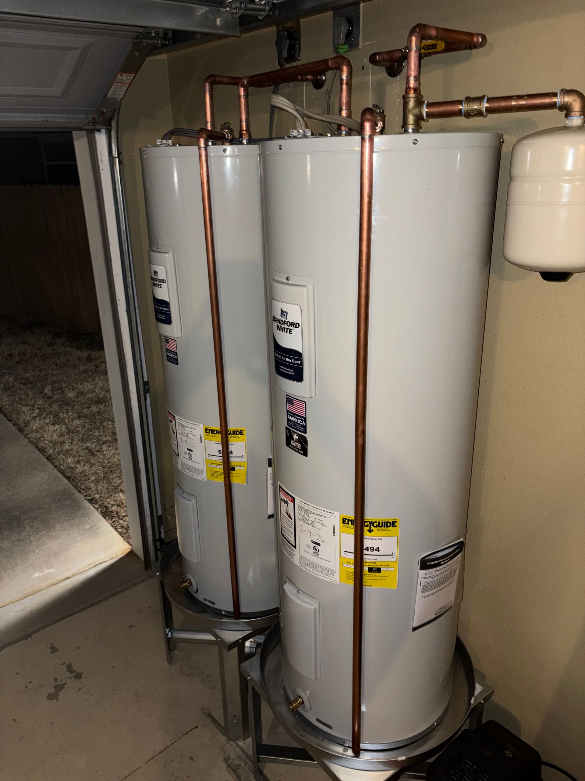 Two white water heaters with copper pipes in a utility closet
