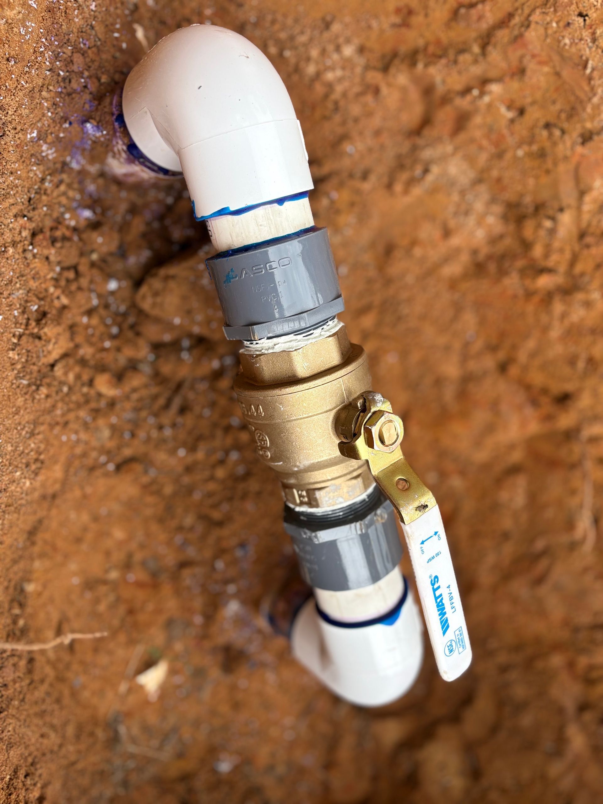 Brass valve on white PVC pipe in a dirt wall cavity, with a small blue tag attached