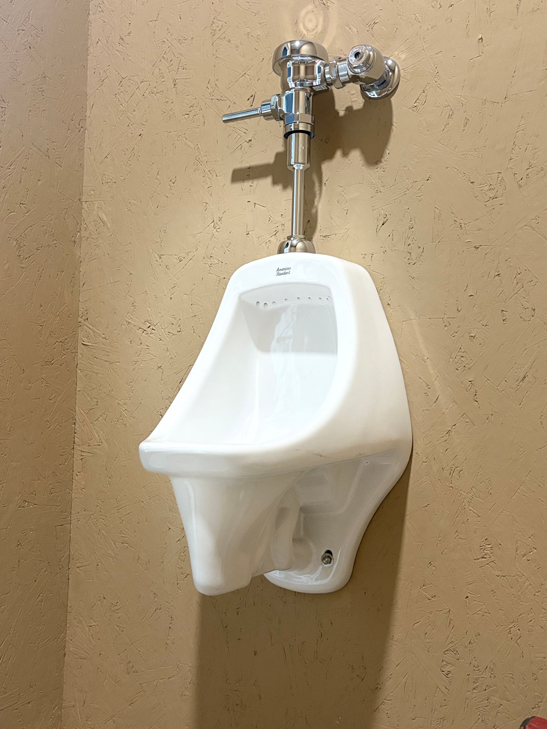 White wall-mounted urinal beneath a chrome flush valve on a tan restroom wall